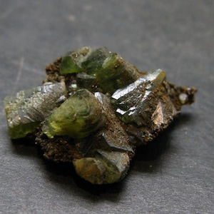 May include: Close-up of a cluster of green crystals with a glassy appearance, set against a dark gray background. The crystals have sharp edges and varying sizes, with a rough, brown matrix.