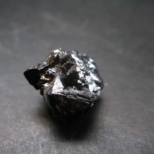 Puede incluir: Primer plano de un espécimen mineral oscuro de forma irregular. La superficie es de un negro profundo con facetas cristalinas reflectantes. El mineral parece estar sobre una superficie gris oscuro.