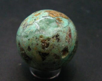 Puede incluir: Una esfera verde, marrón y blanca con una superficie rugosa y texturizada. La esfera tiene una grieta que revela un interior blanco y cristalino.