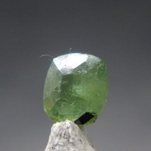 May include: A faceted, light green gemstone with a slightly rounded rectangular shape. The gem is set on a small, light-colored rock, with a dark gray background. The gem has a reflective surface.