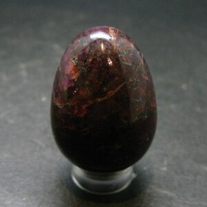 May include: A polished, egg-shaped gemstone featuring a deep, dark burgundy and maroon colour palette with visible internal veining and natural mineral inclusions. The surface is smooth and reflective, showcasing a glossy finish throughout the stone.
