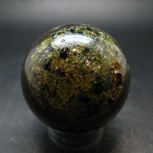 May include: A polished spherical stone featuring a mix of deep forest green, olive, and dark brown mineral inclusions. The surface displays a glossy, reflective finish with visible crystalline textures and dark, opaque patches throughout the orb.