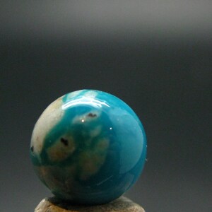 May include: A polished, spherical gemstone featuring a vibrant turquoise blue color with natural white and tan inclusions and small dark brown spots, showcasing a smooth, glossy surface texture and a rounded, orb-like shape.