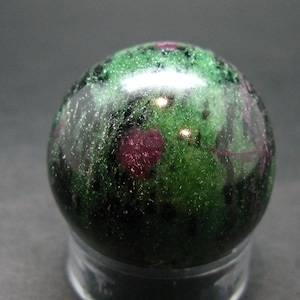 Può includere: Una sfera di pietra preziosa lucidata con una miscela di colori verdi e rosso scuro. La sfera è appoggiata su una base trasparente e rotonda. La superficie della sfera ha un aspetto macchiettato.