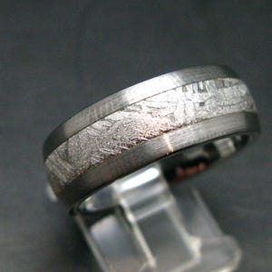 May include: A metallic ring featuring a central inlay with a textured, crystalline pattern. The band has a brushed dark grey finish on the edges, contrasting with the lighter, intricate, and geometric surface of the centre inlay section.