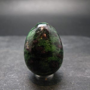 May include: A polished, egg-shaped gemstone with a mix of deep green and dark red hues. The stone has a glossy finish and is set against a neutral background. The gemstone is a decorative item.