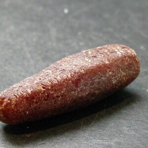 May include: A reddish-brown, oblong gemstone with a textured surface. The stone has a rounded, tapered shape, and appears to be polished. The gemstone is lying on a dark grey surface.