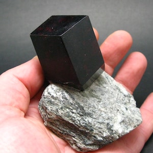 Pode incluir: Um espécime mineral preto escuro, retangular e polido, em cima de uma rocha áspera cinza e branca. O mineral preto tem uma superfície brilhante com sutis inclusões vermelhas. A rocha tem uma textura em camadas.