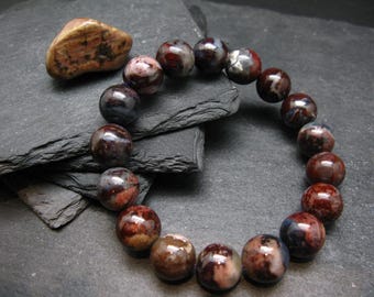 May include: A beaded bracelet featuring large, round beads in shades of red, brown, and blue. The beads have a marbled appearance. A small, polished stone in shades of brown and tan sits nearby. The bracelet is displayed on a dark gray surface.