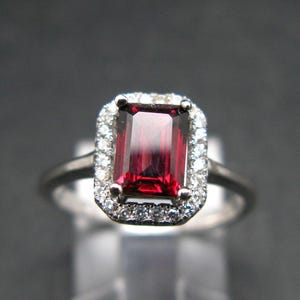 Peut inclure: Une bague en argent avec une grande pierre rouge de taille émeraude entourée d'un halo de petites pierres blanches.