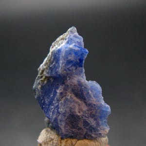 May include: A close-up of a rough, blue mineral specimen. The stone has a crystalline structure with a deep blue color and some lighter, gray-brown areas. The mineral is set against a blurred, dark background.