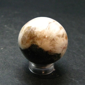 Peut inclure: Une sphère décorative avec une surface polie, présentant un mélange de blanc, de marron et de noir dans un motif distinctif. La sphère est présentée sur un support circulaire transparent, mettant en valeur son design complexe. Diamètre d'environ 3 cm.