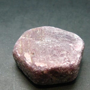 Peut inclure: Une pierre arrondie, rouge-violet, avec une texture légèrement rugueuse. La pierre a une forme naturelle et organique et semble être un spécimen minéral. La couleur est un mélange de rouge profond et de teintes rosées plus claires, avec des taches blanches.
