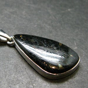 May include: A teardrop-shaped pendant featuring a polished, dark, mottled black and grey stone set in a smooth silver-toned metal bezel. The pendant includes a silver-toned metal bail at the top for attachment to a chain or cord.