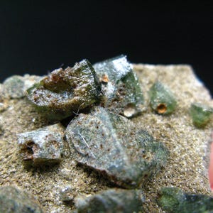 May include: A close-up of a cluster of green and gray crystals embedded in a rough, brown rock. The crystals have a glassy, translucent appearance.