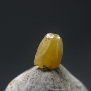 May include: A faceted, golden-yellow gemstone, possibly a topaz or citrine, rests on a light-colored, textured surface. The gem's cut reveals multiple facets, catching the light. The background is a dark, neutral tone, emphasizing the gem's color and form.