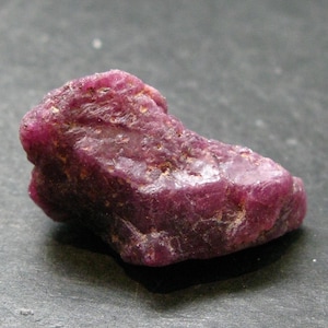Puede incluir: Una gema de rubí en bruto sin cortar. El rubí es de un color rojo intenso y rico con una superficie ligeramente irregular. La gema se encuentra sobre un fondo gris oscuro, resaltando su belleza natural. Este rubí en bruto es un hermoso ejemplo de formaciones minerales naturales.