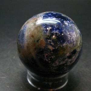 May include: A polished spherical stone featuring a deep blue base color with mottled patches of white, grey, and earthy brown mineral inclusions. The surface of the sphere is smooth and reflective, showcasing a natural, varied pattern throughout the orb.