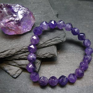 May include: A faceted amethyst bracelet and a raw amethyst crystal. The bracelet has round, deep purple beads. The raw amethyst crystal is a lighter shade of purple. The items are displayed on a dark grey, textured surface.