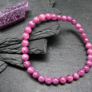 Op de afbeelding: Een roze edelsteenarmband met ronde kralen wordt getoond naast een ruw geslepen, paarse edelsteen. De armband is gemaakt van kleine, gepolijste kralen. De edelstenen zijn geplaatst tegen een donkere, getextureerde achtergrond.