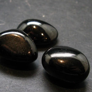 May include: Three polished, black obsidian stones. Each stone has a smooth, rounded shape with a high-gloss finish, reflecting light. The stones are arranged on a dark surface, creating a contrast that highlights their color and texture.