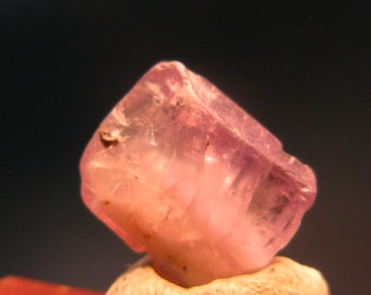 May include: A close-up of a translucent, faceted, pink gemstone. The gem has a rough, natural texture and is set against a dark background. The stone is a light pink color with some areas appearing more opaque.