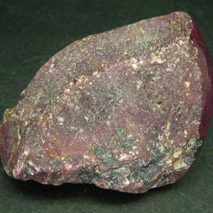 May include: A rough, dark red ruby gemstone with a slightly polished surface. The ruby has a natural, irregular shape and is a deep, rich red colour.