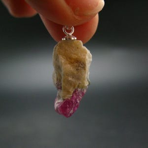 May include: A raw ruby pendant with a silver bail. The ruby is a deep red color and has a rough, natural texture. The pendant is approximately 1 inch in length.