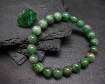 May include: A green beaded bracelet with a matching green gemstone. The bracelet is made of round, polished beads in varying shades of green. The gemstone is a rough, irregular shape. The items are displayed on a dark, textured surface.