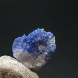 May include: A close-up of a small, rough-textured, blue mineral specimen. The mineral has a crystalline structure and is resting on a light-colored, rounded rock against a dark background.