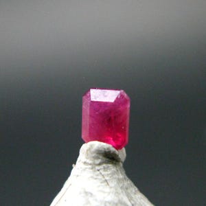 May include: A vibrant, deep pink, emerald-cut gemstone with a faceted surface that reflects light. The stone is positioned atop a textured, light-colored, conical base, showcasing its translucent quality and sharp, geometric edges against a dark background.
