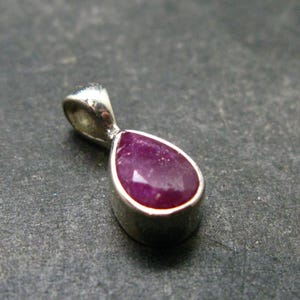 May include: A teardrop-shaped pendant featuring a deep pinkish-red gemstone set in a smooth, polished silver-toned metal bezel. The pendant includes a small, rounded silver-toned bail at the top for attachment to a chain or cord.