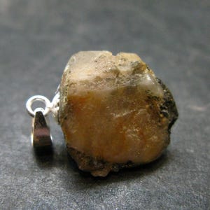 May include: A raw, unpolished tan and brown mineral stone pendant featuring a silver-toned metal bail attached to the side. The stone has a rough, irregular texture with translucent areas and dark inclusions, showcasing a natural, uncut appearance.
