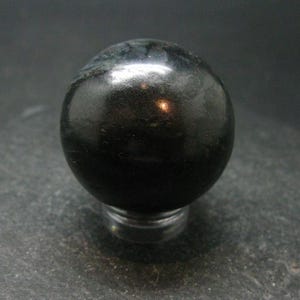 Puede incluir: Una esfera de obsidiana negra lisa, pulida a alto brillo. La esfera descansa sobre una superficie gris oscuro.