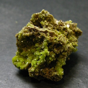 May include: A raw mineral specimen featuring a cluster of vibrant lime green crystals. The structure is irregular and porous, with a mix of bright green crystalline formations and darker, earthy brown textured areas throughout the rock surface.