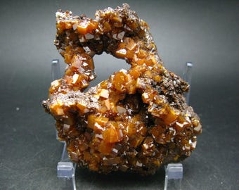 Könnte beinhalten: Ein Cluster aus orange-braunen Kristallen auf einer dunkelbraunen Matrix. Die Kristalle sind in einem Cluster angeordnet, wobei einige der Kristalle einen Hohlraum in der Mitte bilden. Die Kristalle sind durchscheinend und haben einen glasartigen Glanz.