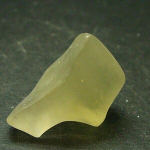 May include: A piece of sea glass, pale yellow in colour, with a smooth, rounded shape. The shard has an irregular form, with a curved edge and a flat, angled surface. The glass has a frosted texture, suggesting it has been smoothed by the sea.