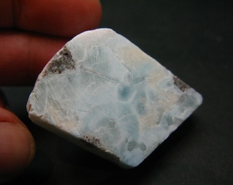 May include: A close-up of a piece of larimar gemstone. The stone has a light blue and white color with darker brown and gray inclusions. The surface has a textured appearance with visible patterns and lines.