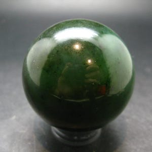 May include: A polished, spherical stone object with a deep forest green colour. The surface is smooth and reflective, showing variations in tone from dark emerald to lighter translucent patches, with a glossy finish that catches the light.