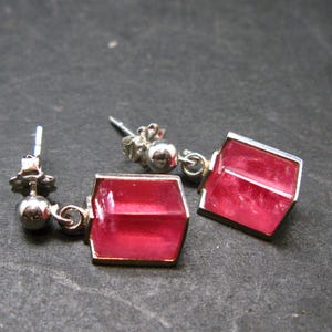May include: A pair of silver-toned earrings with a geometric design. Each earring features a vibrant, rectangular, pink gemstone set within a silver frame. The earrings have a post-and-ball closure.