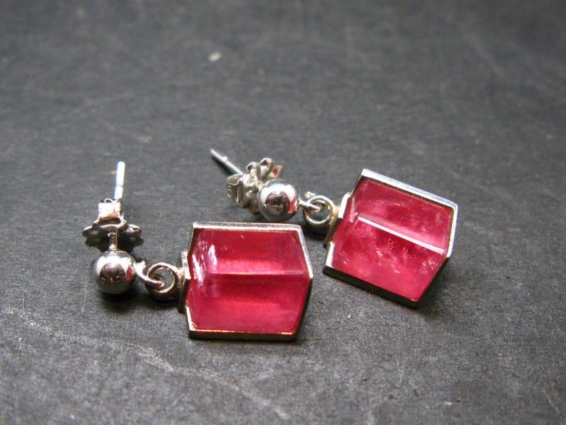 May include: A pair of silver-toned earrings with a geometric design. Each earring features a vibrant, rectangular, pink gemstone set within a silver frame. The earrings have a post-and-ball closure.
