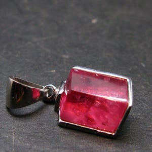 Puede incluir: Un colgante de plata con una gema roja oscura, rectangular y facetada. La gema est&aacute; engastada en un bisel de plata. El colgante tiene una fianza de plata para una cadena. Fondo oscuro y texturizado.