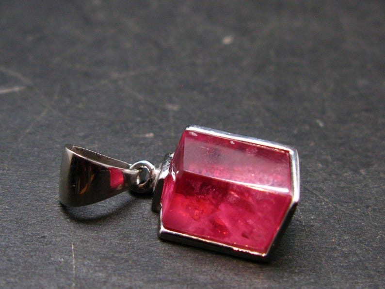 Puede incluir: Un colgante de plata con una gema roja oscura, rectangular y facetada. La gema est&aacute; engastada en un bisel de plata. El colgante tiene una fianza de plata para una cadena. Fondo oscuro y texturizado.