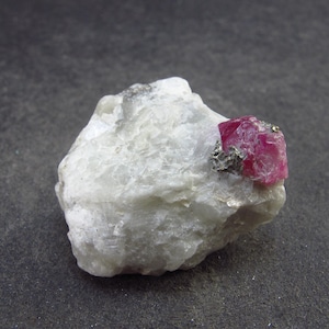 May include: A small, red ruby crystal embedded in a white, opaque matrix. The ruby is a deep red colour and has a sharp, well-defined crystal shape.