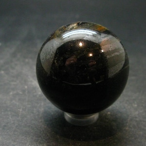 May include: A polished, spherical stone with a deep black base colour, featuring subtle inclusions of brown and metallic grey mineral deposits on its upper surface. The smooth, reflective surface captures light, highlighting its rounded, orb-like shape.