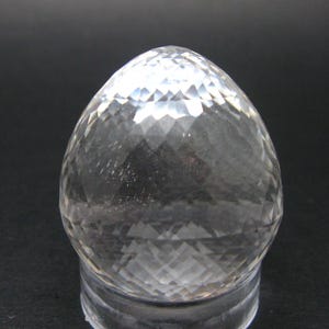May include: A clear, faceted crystal egg-shaped ornament. The crystal is transparent, allowing light to pass through and creating a sparkling effect. The egg sits on a clear, round base, and the background is dark.