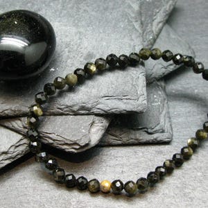May include: A beaded bracelet composed of small, faceted black and gold-sheen obsidian beads arranged in a circular pattern, featuring a single, distinct, round, light-coloured speckled bead positioned near the bottom of the strand.