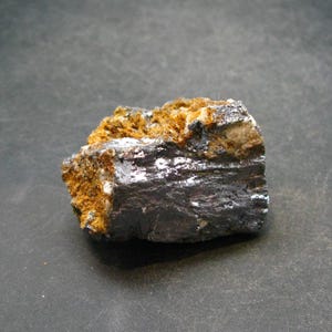 May include: A dark gray, metallic mineral specimen with a rough, textured surface. The mineral is embedded in a matrix of light brown and white material.