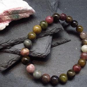 Puede incluir: Un brazalete de cuentas con una variedad de piedras preciosas en tonos tierra, incluyendo tonos de verde, marrón, rosa y gris. El brazalete se exhibe sobre una superficie gris oscuro, con una piedra blanca y rosa en el fondo.