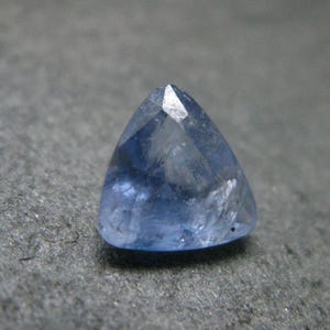 May include: A faceted, triangular-shaped blue gemstone with a translucent appearance and visible internal inclusions. The stone features a polished surface with multiple geometric cuts that reflect light across its light blue and violet-toned facets.